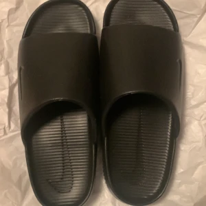 Svarta slides från Nike - Säljer ett par svarta slides från Nike med enkel och stilren design. De har en bred rem över foten och räfflad sula för extra grepp. Perfekta att glida runt i när det är varmt ute.