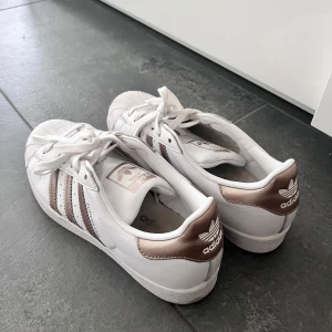 Adidas Superstar med bronsdetaljer - Säljer ett par klassiska Adidas Superstar sneakers i vitt skinn med bronsfärgade ränder och häl. Ikonisk snäcktå och platt sula. Perfekta för dig som vill ha en stilren och sportig look med en twist.