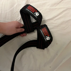 Svarta lifting straps från Gorilla Wear - Säljer 3 svarta straps från Gorilla Wear med röda sömmar. Dom har broderad logga och är gjort för styrketräning. Perfekt för gymmet när du vill ha extra stöd vid tunga lyft.