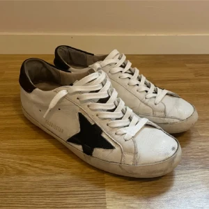 Vita  Golden Goose - Säljer ett par vita sneakers från Golden Goose med svart stjärndetalj på sidan och svarta detaljer vid hälen. Skorna har vita snören och platt sula. Perfekta för en avslappnad och trendig stil. Använda skor men fortfarande riktigt feta TAR BYTEN OCKSÅ