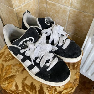 Svarta Adidas Campus sneakers med vita ränder - Säljer ett par klassiska Adidas Campus sneakers i svart mocka med vita ränder och vit sko snören. De är använda ett antal gånger men köpt nya och de kommer inte till användning. Där bak har den en defekt för en hund bet i skon där bak men funkar fortfarande som vanligt och är ett par sköna sneakers, kom med bud!!