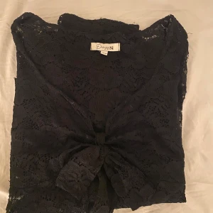 Svart spetsblus med knyt från Design by Si - Säljer en svart blus i spets från Design by Si. Blusen har v-ringning, knyt framtill och trekvartsärm. Perfekt för dig som gillar detaljer och vill ha en feminin look. Spetsmaterialet ger en elegant känsla. Den har endast använts 1 gång.
