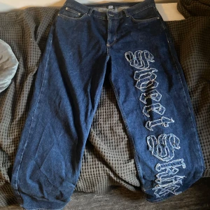 Blå jeans med broderad text - Säljer ett par baggy blå jeans med stor broderad text 'Sweet Skillz' längs ena benet. Byxorna har klassisk femficksdesign och raka ben. Perfekta för dig som vill sticka ut med en unik detalj på dina jeans. Det är lite fransigt längst ner på dom men annars är dom i bra skick. 