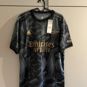 Arsenal bortatröja 2022/23 från Adidas - Säljer en Arsenal bortatröja från Adidas i svart och guld med geometriskt mönster. Tröjan har korta ärmar, guldfärgade loggor och sponsortryck. Materialet är lätt och andas, perfekt för match eller träning.