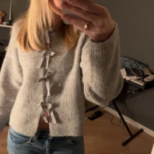 Grå stickad kofta med rosetter i storlek S - Superfin grå stickad kofta med dekorativa rosetter framtill. Koftan har lång ärm och en croppad passform som passar perfekt till jeans eller kjol. Mysig och trendig för kyligare dagar.