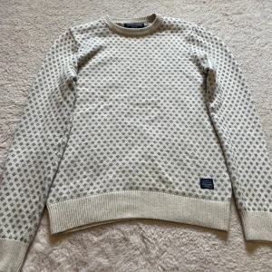 Vit och gråmönstrad stickad tröja från Bondelid - Säljer en vit stickad tröja från Bondelid med grått småmönster över hela tröjan. Tröjan har rund halsringning, långa ärmar och ribbade muddar vid ärmslut och nederkant. Perfekt för kyligare dagar!