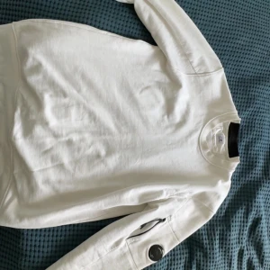 Vit sweatshirt från C.P. Company - Säljer en stilren vit sweatshirt från C.P. Company med rund hals och lång ärm. Tröjan har en ikonisk linsdetalj på ena ärmen och en praktisk ficka med dragkedja. Perfekt för dig som gillar streetwear och snygga detaljer.