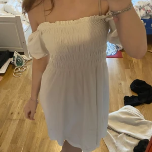 Vit klänning med puffärm - Söt vit klänning med smock upptill. Klänningen har korta puffärmar och är tillverkad i ett lätt tyg, perfekt för varma dagar. Går att ha både off shoulder och över axlarna.