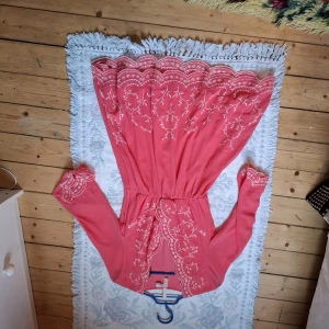 Korallrosa långklänning med broderade detaljer - Superfin korallrosa långklänning med vackra broderade mönster och volangkant upptill. Klänningen har lång ärm och markerad midja, perfekt för sommarens tillfällen. Broderierna ger en romantisk känsla och färgen är riktigt härlig.