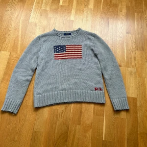 Polo Ralph Lauren USA - Säljer en grå stickad tröja från Polo Ralph Lauren med en stor amerikansk flagga framtill och RL-broderi vid nederkanten. Tröjan har rund hals och långa ärmar. Perfekt för dig som gillar klassisk och stilren design. Riktigt snyggt röda detaljer parfeckt för sommar kvällar tveka inte ens på att hör av sig😊