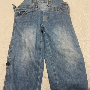 Blå jeans hängselbyxor från Okaou - Säljer ett par klassiska blå jeans hängselbyxor från Okaou, La Redoute Creation för bebisar 2-3 år. Byxorna har knappar framtill och på sidorna, samt fickor både fram och bak. Perfekta för en avslappnad och cool stil.
