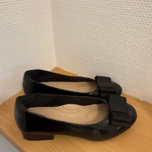 Helt nya ballerinaskor - Stilrena svarta ballerinaskor i glansigt material med en stor dekorativ rosett framtill. Skorna har låg klack och rund tå, perfekta för en klassisk look. De har använts vid ett tillfälle så därav defekter. Köptes för 800 men säljer för 450❤️
