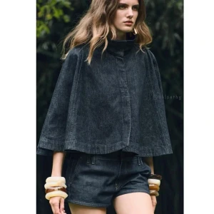 Svart jeanscape med hög krage - Säljer en svart cape i jeansmaterial med hög krage och vid passform. Jackan har en minimalistisk design utan synliga knappar eller dragkedja framtill. Perfekt för dig som vill ha en unik och trendig look.