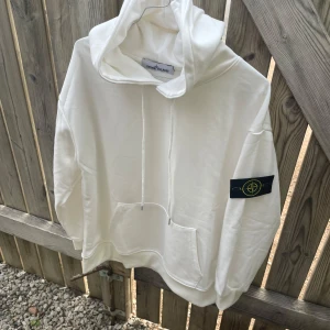Vit hoodie från Stone Island - Säljer en stilren vit hoodie från Stone Island med klassisk patch på vänster ärm. Tröjan har huva med dragsko och en stor magficka framtill. Perfekt för dig som gillar streetwear och vill ha något enkelt men snyggt. Storlek s men passar en xs med.