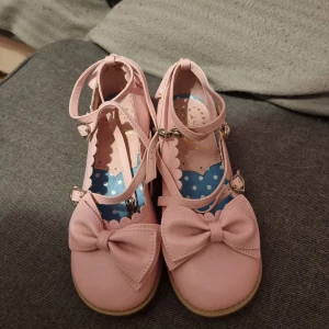 Rosa ballerinaskor med rosett och remmar - Säljer ett par söta rosa ballerinaskor med stora rosetter framtill och dubbla remmar med spänne med hjärtan. Skorna har låg klack och rund tå, samt dekorativa detaljer även baktill. Passar kawaii, fairykei, lolita stilar. Köpte för en cosplay 9 år sedan men de är aldrig använda och förtjänar att komma till ett hem där de används. Kostade 500 kr nya pris.