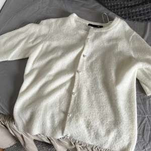 Vit stickad kofta med pärlknappar - Mysig vit stickad kofta från H&M med korta ärmar och pärlknappar framtill. Perfekt att slänga på sig över en topp eller klänning. Ribbad kant vid hals, ärmslut och nederkant.