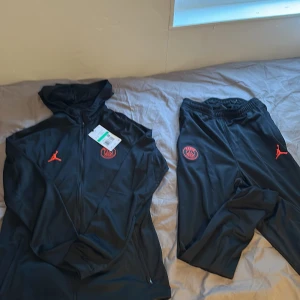 Svart PSG x Jordan träningsset med röd logga - Säljer ett svart träningsset från PSG x Jordan i storlek XL. Setet består av en hoodie med dragkedja och matchande byxor, båda med röda PSG-loggor och Jordan-detaljer. Perfekt för fotbollsträning eller streetwear.