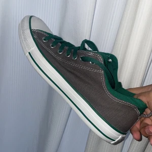 Grå och gröna Converse sneakers - Säljer ett par unika Converse sneakers i grå canvas med gröna detaljer och vita gummisulor. Skorna har snörning och rund tå, perfekt för dig som gillar en stilren men färgglad look. De är i mycket bra skick och det finns knappt några tecken på användning.