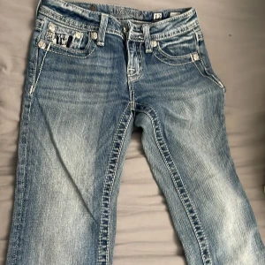 Blå bootcut jeans från Miss Me - Säljer ett par blå bootcut jeans från Miss Me med slitningar och dekorativa bakfickor med broderade detaljer och nitar. Jeansen har normal passform och klassisk femficksdesign. Perfekta för dig som gillar snygga detaljer och unik stil. Midjemått: 30-31 cm innerbenslängd: 68-69cm
