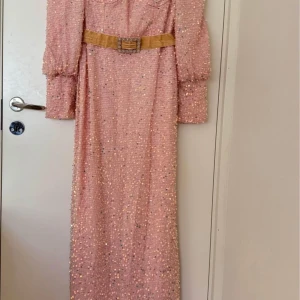 Ljusrosa glittrig långklänning med puffärmar och bälte - Elegant ljusrosa långklänning med glittriga paljetter, markerad midja med dekorativt bälte och långa puffärmar. Klänningen har fyrkantig ringning och figurnära passform som framhäver siluetten. Perfekt för bal eller fest.