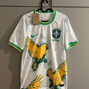 Vit Brasilien fotbollströja från Nike - Säljer en vit Brasilien fotbollströja från Nike med gröna och gula detaljer. Tröjan har korta ärmar, grönt och gult runt kragen, samt ett stort tryck av gula fåglar och Brasilien-emblemet på bröstet. Materialet är Dri-FIT för bra andningsförmåga.