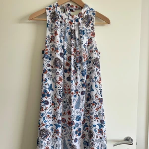 Vit blommig ärmlös klänning med knytband - Säljer en vit ärmlös klänning med färgglatt blommönster i blått, rött och svart. Klänningen har en hög hals med knytband och ett luftigt, rakt fall. Perfekt för varma dagar och somriga tillfällen.