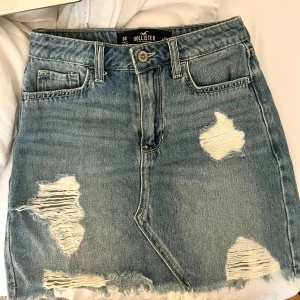 Blå jeanskjol från Hollister - Säljer en blå jeanskjol från Hollister med slitna detaljer och fransad nederkant. Kjolen har klassisk femficksdesign och knappgylf. Perfekt för en avslappnad stil.
