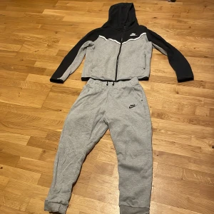 Full Tec fleece från Nike  - Säljer en snygg hoodie från Nike i grått och svart med dragkedja och huva. Tröjan har långa ärmar och en vit Nike-logga på bröstet. Perfekt för träning eller chill. Matchande gråa mjukisbyxor medföljer.