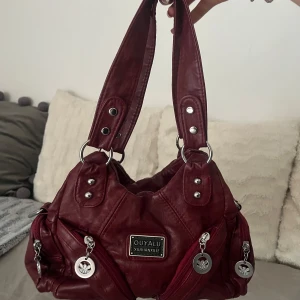 Vinröd axelväska - Säljer en vinröd axelväska från OUYALU med flera dragkedjor och silvriga detaljer. Väskan har både korta och lång justerbar rem, samt dekorativa hängsmycken. Perfekt för dig som vill ha en snygg och praktisk accessoar.