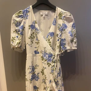 Blommig omlottklänning från & other Stories - Söt vit omlottklänning med blommönster. Klänningen har puffiga korta ärmar, volang nertill och knyt i midjan. Perfekt för sommarens tillfällen.