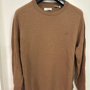 Brun stickad tröja i merinoull från Gant - Säljer en brun stickad tröja från Gant i 100% merinoull. Tröjan har ett diskret mönster, rund halsringning och långa ärmar. Perfekt för dig som gillar stilrena och bekväma plagg.