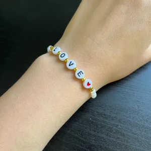 Armband med bokstavspärlor LOVE🩷 - Fint armband med vita runda pärlor som bildar ordet LOVE och ett rött hjärta. Mellan bokstäverna sitter små guldfärgade pärlor. Perfekt accessoar för att sprida kärlek. 🩷