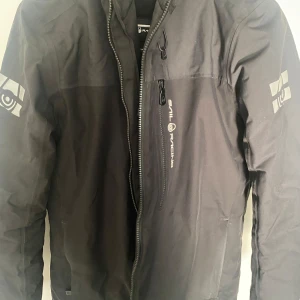 Svart vindjacka från Sail Racing - Säljer en svart vindjacka från Sail Racing med dragkedja framtill och logga på bröstet samt ärmarna. Jackan har huva och en sportig look, perfekt för blåsiga dagar.