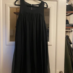 Svart ärmlös klänning med dragkedja - Svart, luftig och ärmlös Rodebjer klänning med rund halsringning och dragkedja i ryggen. Klänningen har ett plisserat fall och är tillverkad i silke som ger en fin siluett. Perfekt för dig som gillar enkel och stilren design.