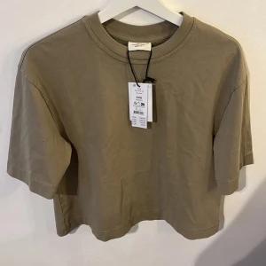 Beige boxy t-shirt från Gina Tricot - Säljer en basic boxy t-shirt i en snygg grön färg från Gina Tricot. T-shirten har en avslappnad passform, korta ärmar och rund halsringning. Perfekt för en enkel och stilren look. Helt ny och oanvänd!