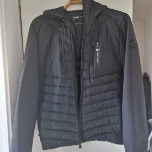Svart pufferjacka från Sail Racing - Säljer en svart pufferjacka från Sail Racing med huva och dragkedja. Jackan har två bröstfickor med dragkedja och Sail Antarctica Expedition-detalj på ärmen. Perfekt för kyliga dagar. 