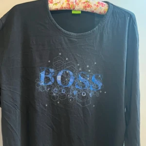 Svart långärmad t-shirt från Hugo Boss x Smiley - Cool svart långärmad t-shirt från Hugo Boss i samarbete med Smiley. Framsidan har ett blått BOSS-tryck med hexagonmönster och baksidan är dekorerad med flera smiley-ansikten med hörlurar. Perfekt för dig som gillar streetwear och unika prints.