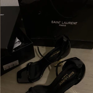 Svarta klackskor från Saint Laurent med YSL-detalj - Säljer ett par svarta klackskor från Saint Laurent med öppen tå och ikonisk YSL-logga i guld som klack. Skorna har en elegant rem runt vristen och är tillverkade i blankt material. Perfekta för dig som vill sticka ut med lyxig design. Köpte de eftersom jag bara skulle ha på de i balen, och nu kommer de inte komma till användning. Så kan säga att det är nytt skick