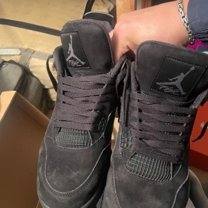 Jordan 4 black cats - Säljer ett par svarta Nike Air Jordans 4 Retro med snygg mocka och klassisk siluett. Skorna har snörning, rund tå och platt sula. Perfekta för dig som vill ha en ikonisk sneaker med stilren look.