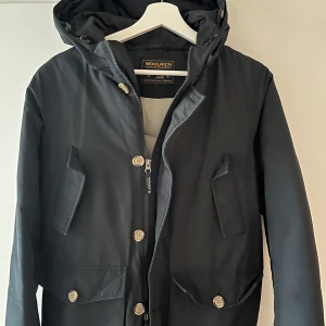 Svart jacka med huva från Woolrich - Säljer min svart/marinblå vinterjacka från woolrich. Den håller värmen otroligt bra och har 2 fodrade fickor upptill. Den är använd 2 vintrar men skicket är bra! Nypris 7300kr