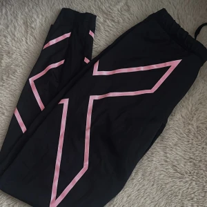 Svarta träningstights från 2XU med rosa detaljer - Svarta träningstights från 2XU med rosa grafiska linjer längs benen. Tightsen har elastisk midja och är tillverkade i ett stretchigt material som passar perfekt för träning.
