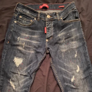 Mörkblå slitna jeans från Dsquared2 - Säljer ett par mörkblå jeans från Dsquared2 med slitningar och små färgstänk. Jeansen har klassisk femficksdesign, knappgylf och röda Dsquared2-detaljer. Snyggt Dsquared2-märke på bakfickan. Perfekta för en trendig streetstil.
