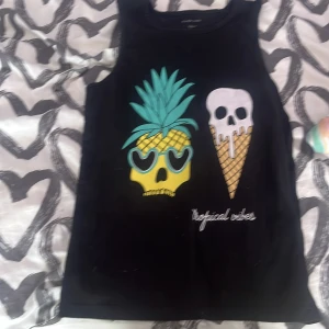 Svart linnetopp med tryck från Ellos - Cool svart linnetopp från Ellos med färgglatt tryck av en ananas-skalle med solglasögon och en glass med döskalle. Texten 'tropical vibes' nedtill. Perfekt för sommarens varma dagar!