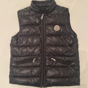 Svart dunväst från Moncler - Snygg svart dunväst från Moncler med hög krage och dragkedja framtill. Västen har två snedställda fickor med dragkedja och Moncler-logga på bröstet. Perfekt för lager-på-lager och stilren look.