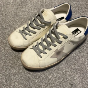 Golden Goose - Golden Goose Superstar sneakers i vitt skinn med grå snörning, grå stjärndetalj på sidan och blå hälkappa. Skorna sitter som 43. PRIS KAN DISKUTERAS. 