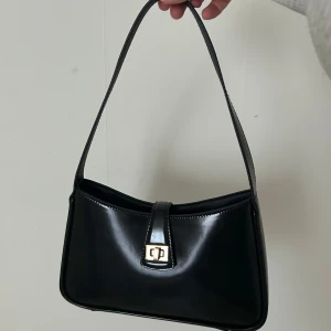 Svart axelväska från H&M - Stilren svart axelväska i glansigt material med guldfärgat spänne framtill. Väskan har en enkel, klassisk form och ett smalt axelband. 