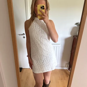 Vit spetsklänning med halter neck - Säljer en söt vit kortklänning med hög hals och ärmlös design. Klänningen har ett blommigt spetsmönster och en lätt A-linjeform. Perfekt för sommaren eller student!!!