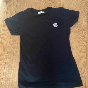 Svart t-shirt från Moncler - Säljer en svart t-shirt från Moncler med liten broderad logga på bröstet. Klassisk passform och korta ärmar, perfekt till vardags. Skön och stilren modell med rund halsringning.
