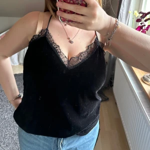Svart topp med spetsdetaljer - Säljer en svart topp med tunna axelband och v-ringning. Toppen har fina spetsdetaljer både fram och bak, där ryggen är helt i spets med ett dekorativt mönster. Perfekt att styla med jeans eller kjol.