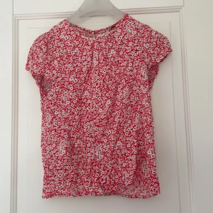 Röd blommig blus från Holly & Whyte - Säljer en söt röd blus med vitt blommönster från Holly & Whyte by Lindex. Blusen har korta ärmar och rund halsringning. Perfekt för att liva upp garderoben med lite färg!